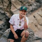 Casual portrait of a smiling man with tattoos sitting on coastal rocks.