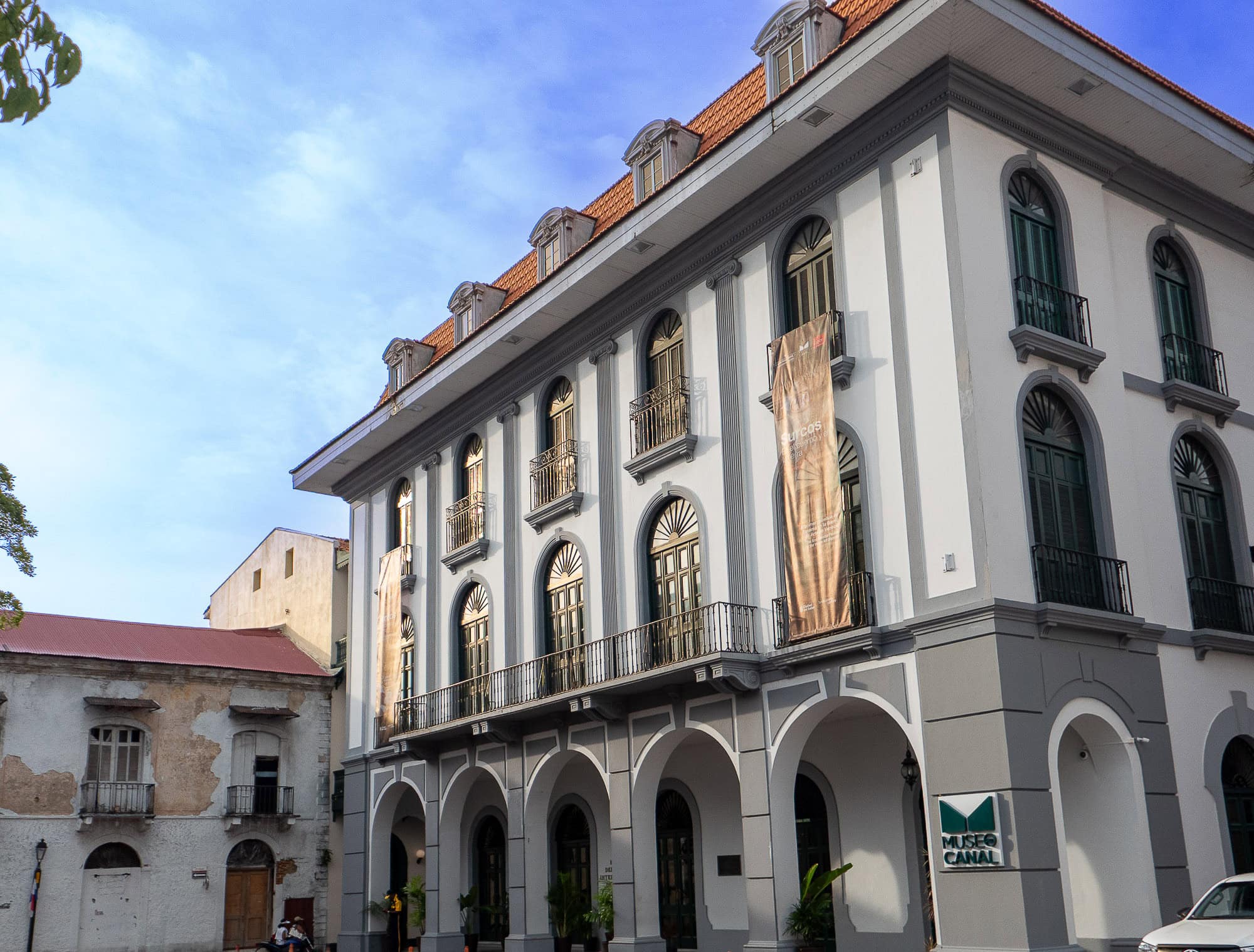 Front façade of Museo del Canal Interoceánico in Casco Viejo, Panama, with neoclassical design, banners, and green shuttered windows under a red-tiled roof.